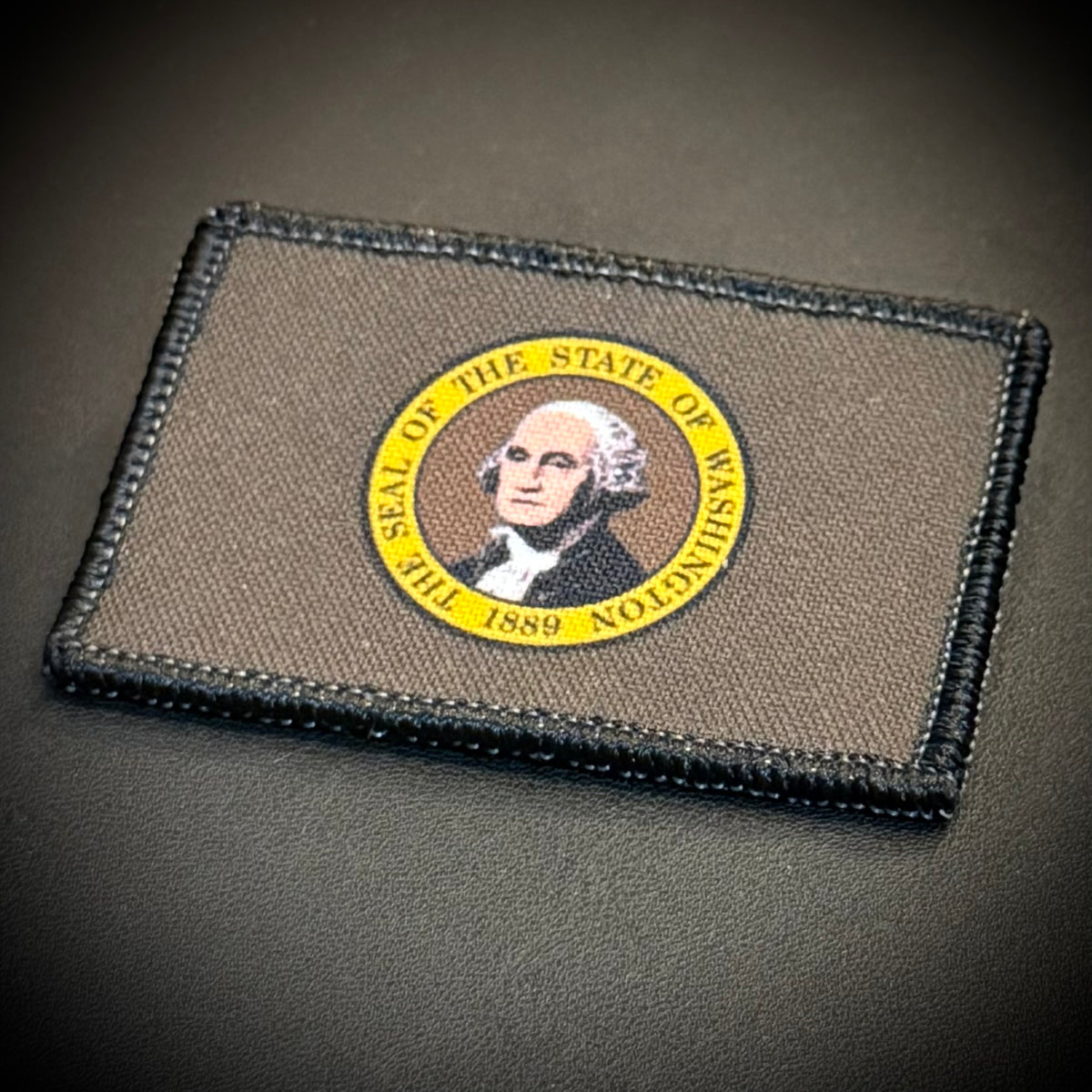 A rectangular tactical patch featuring the seal of the State of Washington, displayed on a dark textured surface.