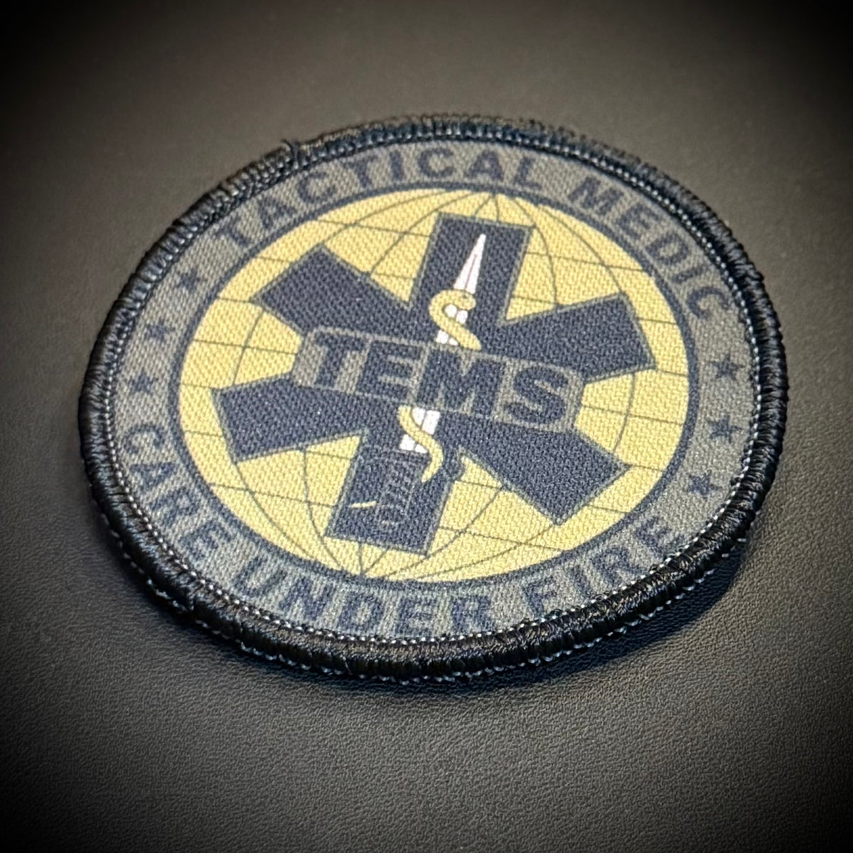 A circular tactical medic care under fire patch with a black border, yellow background, and a dark olive Star of Life symbol featuring a snake and sword.