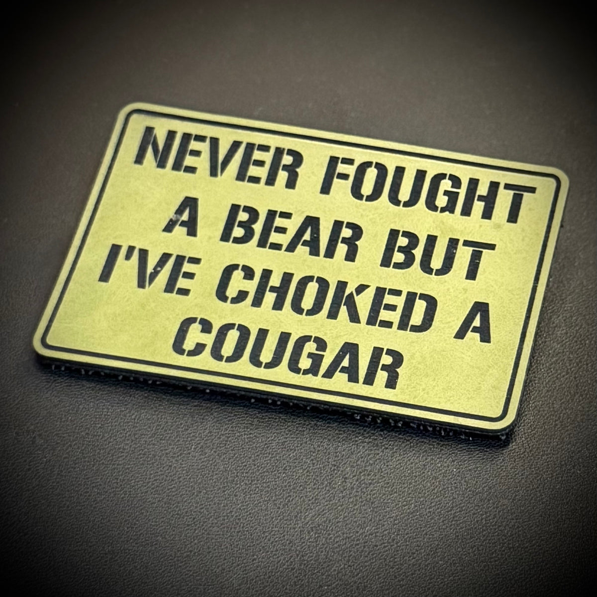I’ve Choked A Cougar laser-engraved synthetic morale patch: a rectangular novelty patch with a pale yellow background and black border, featuring the text 'NEVER FOUGHT A BEAR BUT I'VE CHOKED A COUGAR' in black stencil-style.
