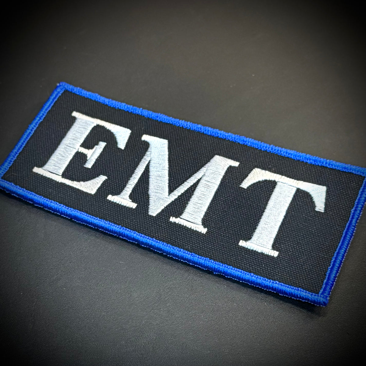 An embroidered rectangular patch with the letters 'EMT' in white on a black background, bordered by a bright blue edge, shown on a dark surface.