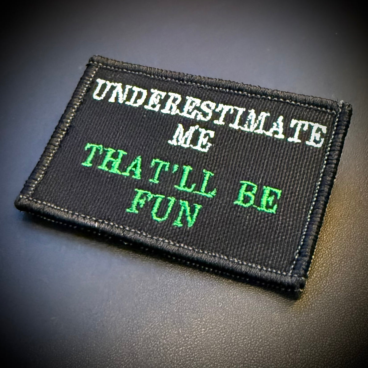 A rectangular black embroidered patch with the text 'UNDERESTIMATE ME THAT'LL BE FUN' in white and green lettering, shown on a dark textured surface.