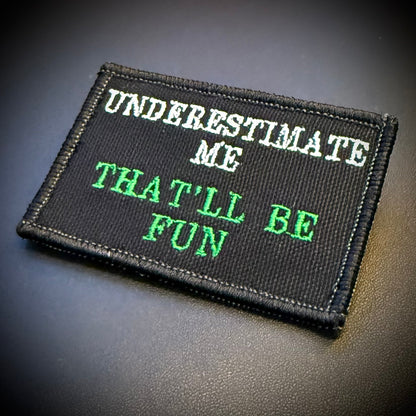 A rectangular black embroidered patch with the text 'UNDERESTIMATE ME THAT'LL BE FUN' in white and green lettering, shown on a dark textured surface.