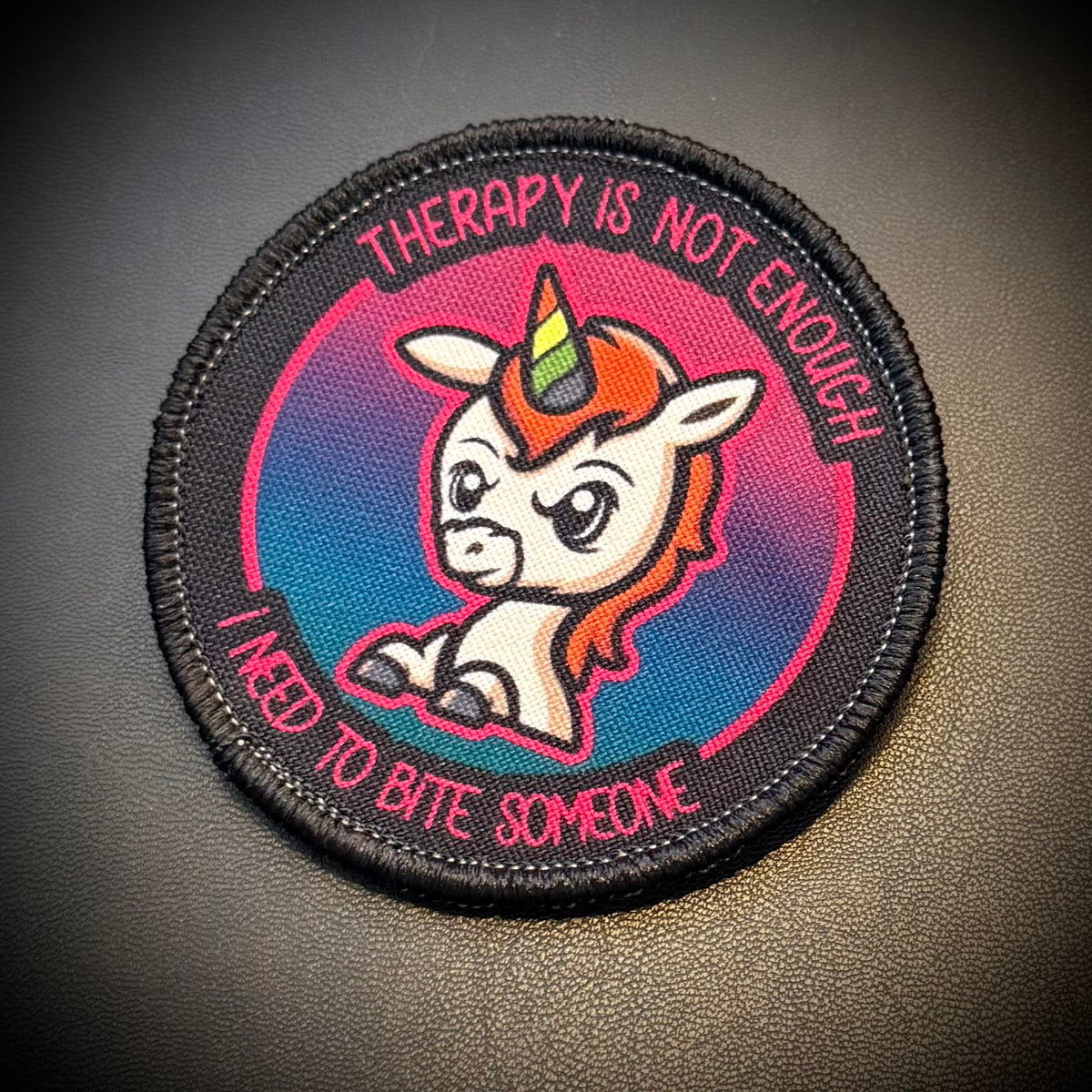 A circular embroidered patch featuring a cartoon unicorn with a rainbow horn, set against a pink-to-blue gradient background with the text 'THERAPY IS NOT ENOUGH' and 'I NEED TO BITE SOMEONE' in pink lettering, shown on a dark textured surface.