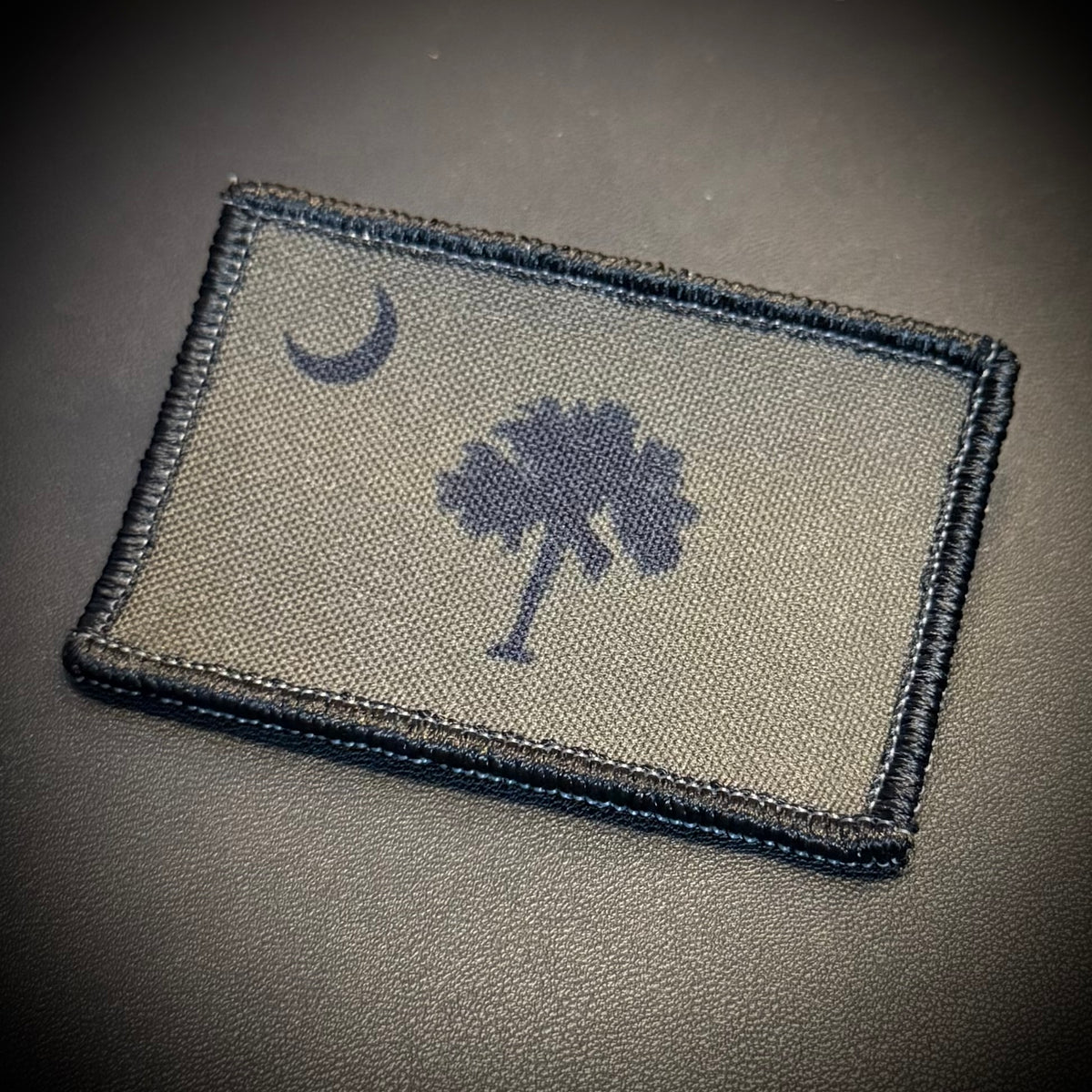 A rectangular embroidered tactical patch featuring the South Carolina state flag design, including a crescent moon and palmetto tree, in dark navy on a khaki background, shown on a dark textured surface.