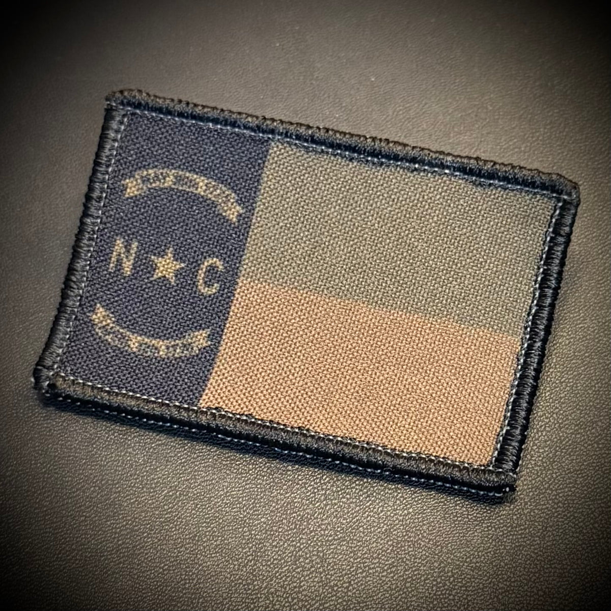 A rectangular embroidered patch featuring the North Carolina state flag design in subdued colors, placed on a dark textured surface. The patch has a dark blue vertical band with a star and the letters 'N C', plus a tan and gray horizontal block layout.