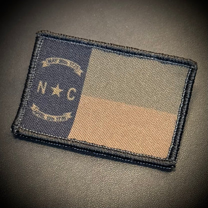 A rectangular embroidered patch featuring the North Carolina state flag design in subdued colors, placed on a dark textured surface. The patch has a dark blue vertical band with a star and the letters 'N C', plus a tan and gray horizontal block layout.