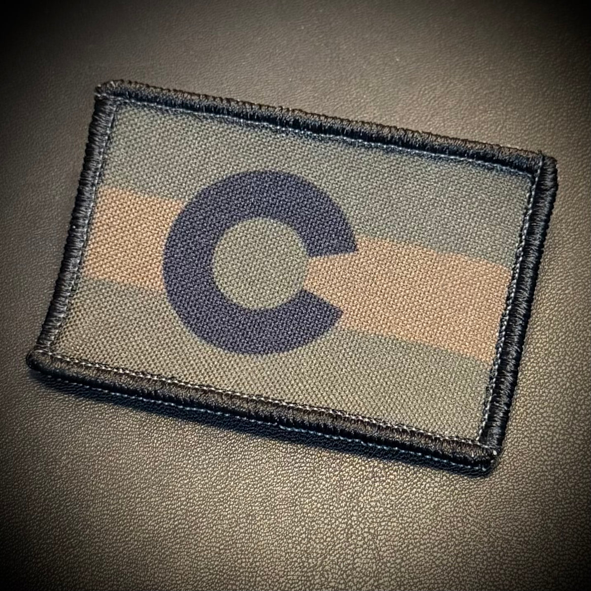 A rectangular embroidered patch featuring a dark blue 'C' symbol over horizontal stripes in muted green, beige, and gray, finished with a dark blue stitched border, shown on a dark textured surface.