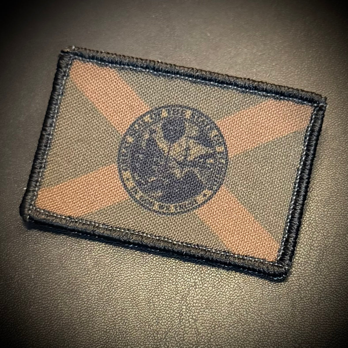 A rectangular embroidered tactical patch featuring a dark navy blue seal in the center, overlaid on a diagonal camouflage pattern of tan and olive green, all enclosed in a black stitched border. The patch is photographed on a dark textured surface.