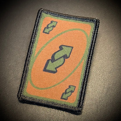 A rectangular tactical patch featuring a black border, an orange background, a green oval, and green double-arrow symbols, displayed on a dark textured surface.