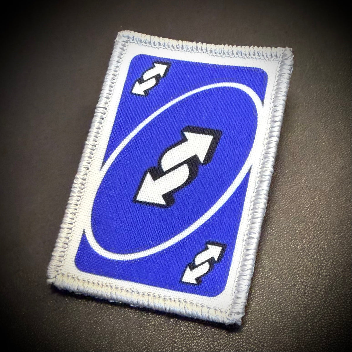 A rectangular embroidered patch featuring a blue background with a white oval, double arrows, and two small white double-arrow symbols in the corners, shown on a dark textured surface.