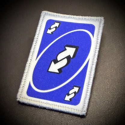 A rectangular embroidered patch featuring a blue background with a white oval, double arrows, and two small white double-arrow symbols in the corners, shown on a dark textured surface.