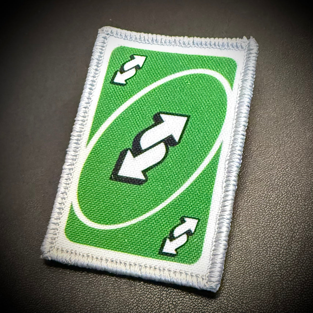 A rectangular embroidered patch designed like a green card with a white border, featuring a double arrow symbol in the center and smaller matching arrows in the corners, shown on a dark textured background.