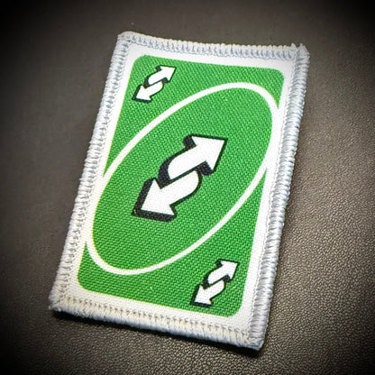 A rectangular embroidered patch designed like a green card with a white border, featuring a double arrow symbol in the center and smaller matching arrows in the corners, shown on a dark textured background.