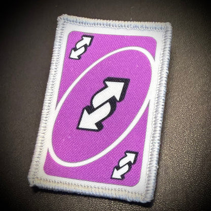 A rectangular embroidered patch designed to look like a purple playing card with white border stitching, featuring a central double-arrow symbol and smaller arrows in the corners, shown on a dark textured background.