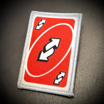 A rectangular embroidered patch featuring a red background with a white oval, double arrows, and small white arrows, finished with a white stitched border, shown on a dark textured surface.