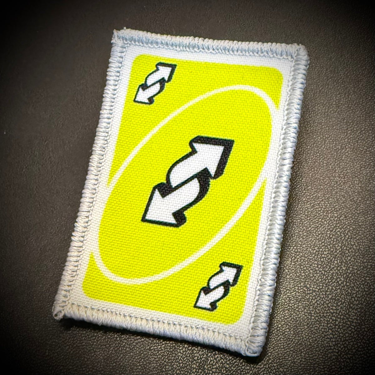A rectangular embroidered patch featuring a bright yellow background with a white oval, black-and-white double arrows in the center, and two smaller matching arrows in the corners, shown on a dark textured surface.