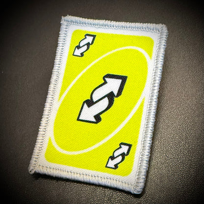 A rectangular embroidered patch featuring a bright yellow background with a white oval, black-and-white double arrows in the center, and two smaller matching arrows in the corners, shown on a dark textured surface.