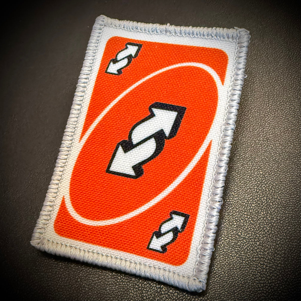 A rectangular embroidered patch featuring an orange and white card design with black and white arrows, placed on a dark textured surface.