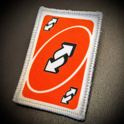 A rectangular embroidered patch featuring an orange and white card design with black and white arrows, placed on a dark textured surface.