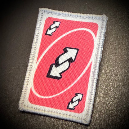 An embroidered patch shaped like a red and white Uno card, featuring the classic double-arrow symbol in black and white, displayed on a dark textured surface.