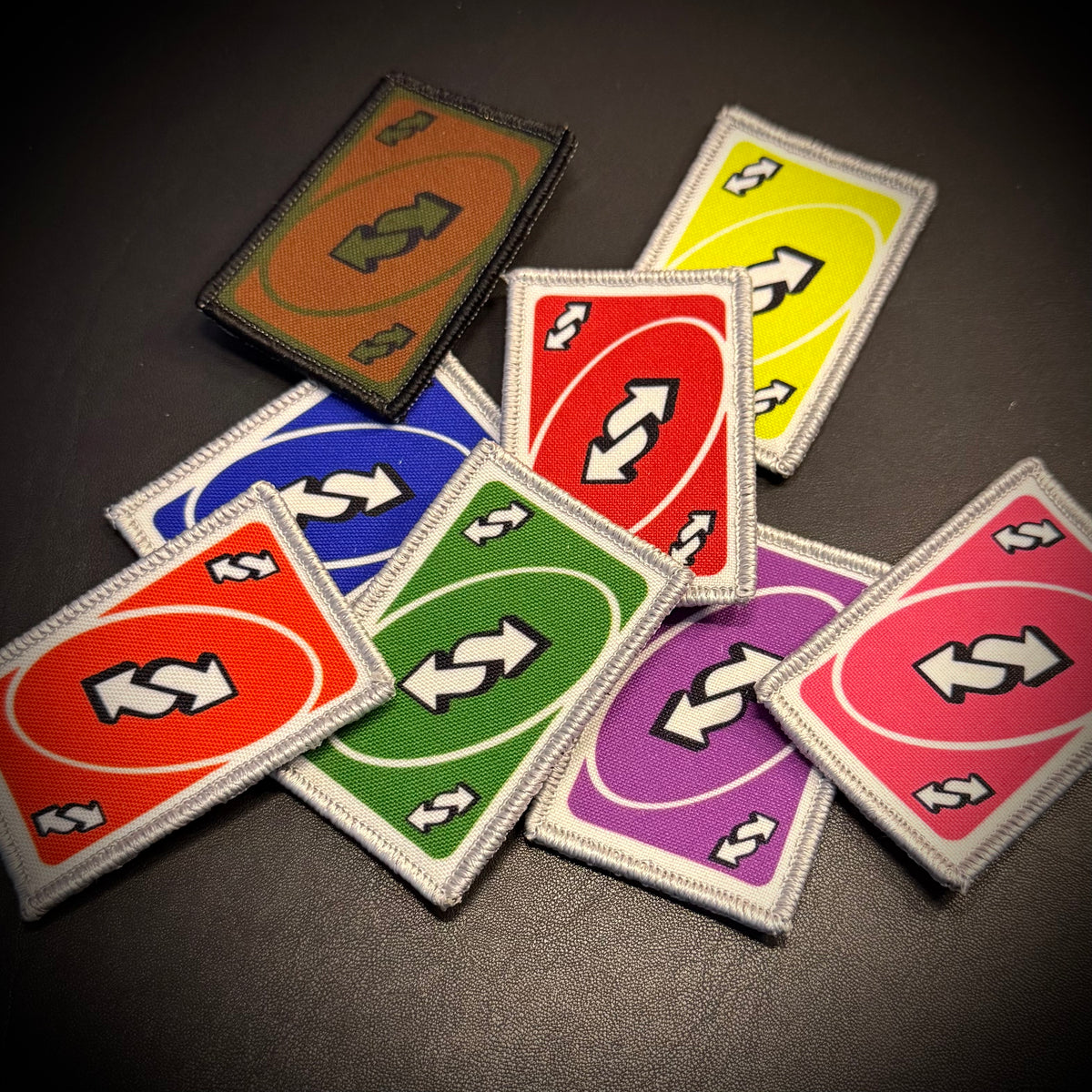 A set of colorful rectangular embroidered patches with white borders, arranged on a dark surface. The patches feature a red-orange oval with two white arrows, a green oval with two white arrows, a blue oval with two white arrows, a yellow oval with two white arrows, a purple oval with two white arrows, and a brown oval with two white arrows.