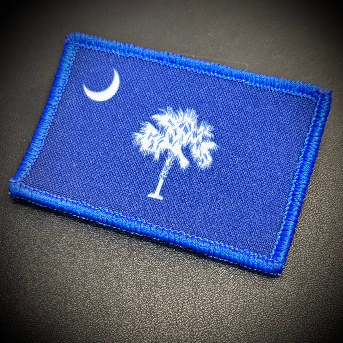 A rectangular embroidered patch featuring the South Carolina state flag design, with a blue field, white crescent moon, and white palmetto tree, bordered by blue stitching on a dark textured background.
