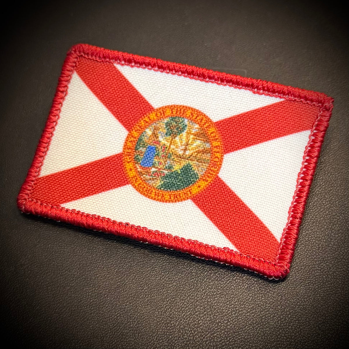 An embroidered rectangular patch of the Florida state flag, featuring a red saltire on a white field with the state seal in the center, photographed on a dark textured surface.