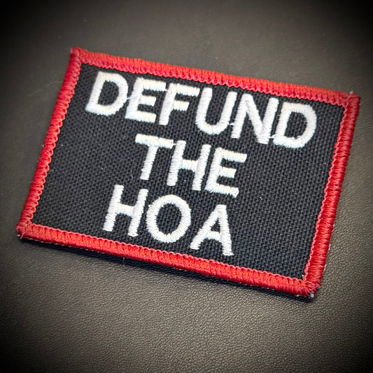 A rectangular embroidered patch with the text 'DEFUND THE HOA' in white lettering on a black background, bordered by a red stitched edge, shown on a dark textured surface.