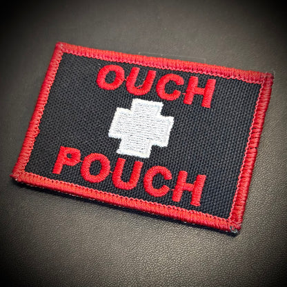 An embroidered rectangular patch with a black background, red border, red text reading 'OUCH POUCH,' and a white cross in the center, photographed on a dark textured surface.