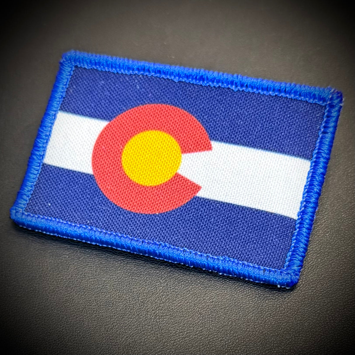 A rectangular embroidered patch of the Colorado state flag with blue border, blue and white horizontal stripes, and a red circle with a yellow center, shown on a dark textured surface.