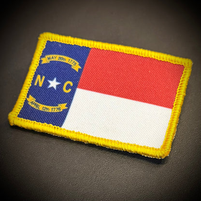 An embroidered rectangular patch of the North Carolina state flag with a yellow stitched border, placed on a dark textured surface.