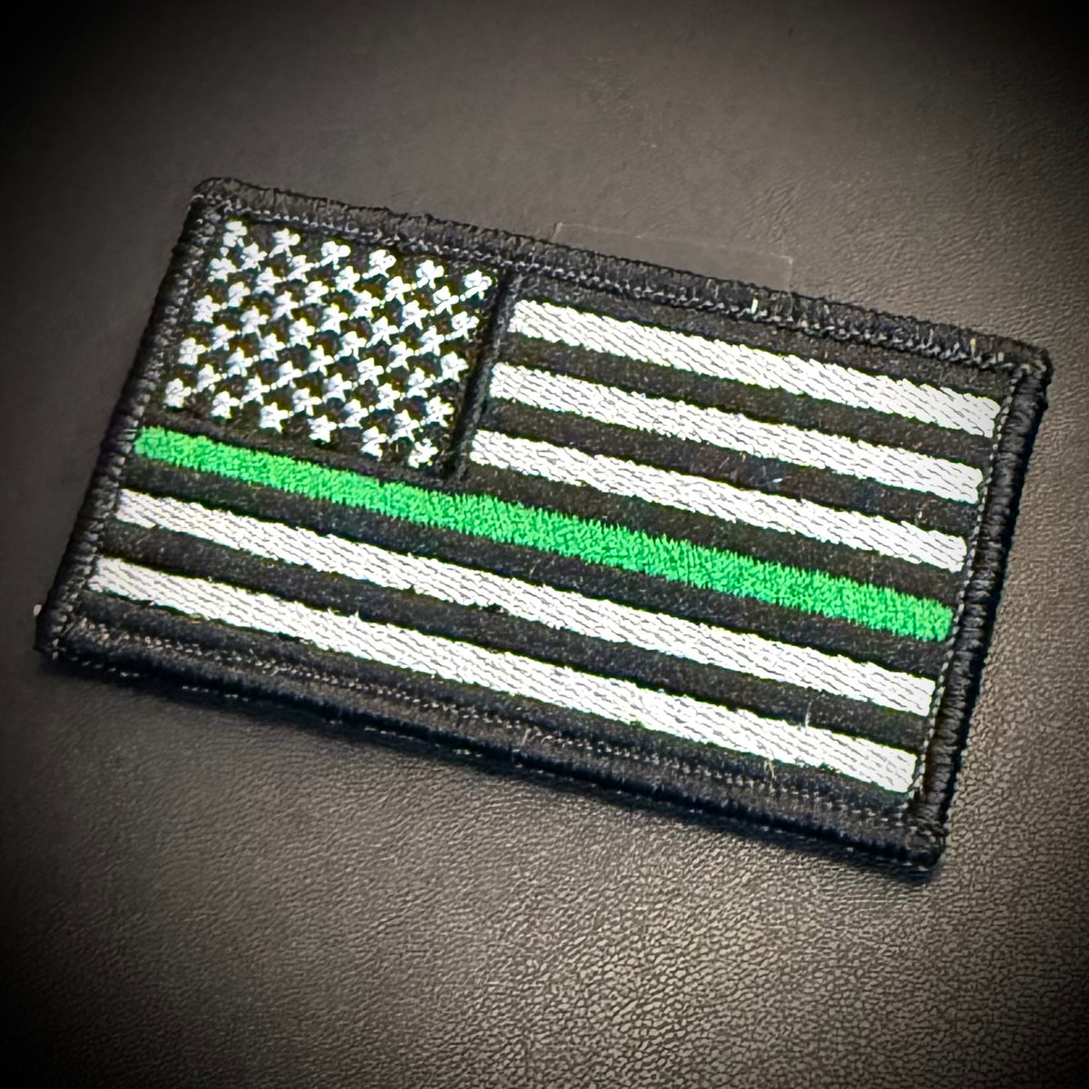 An embroidered American flag patch with a thin green line, shown on a dark textured surface.