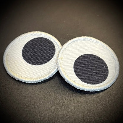Two round white fabric patches with black circular centers, resembling cartoon eyes, displayed on a dark textured surface.