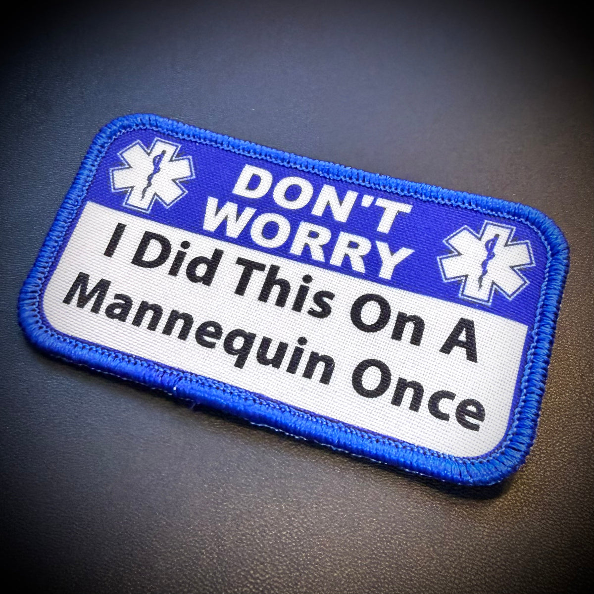 A rectangular embroidered patch with a blue border and top section reading 'DON'T WORRY' with two white medical-style symbols, and a white lower section reading 'I Did This On A Mannequin Once' in black text, shown on a dark textured background.