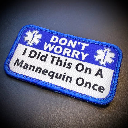 A rectangular embroidered patch with a blue border and top section reading 'DON'T WORRY' with two white medical-style symbols, and a white lower section reading 'I Did This On A Mannequin Once' in black text, shown on a dark textured background.