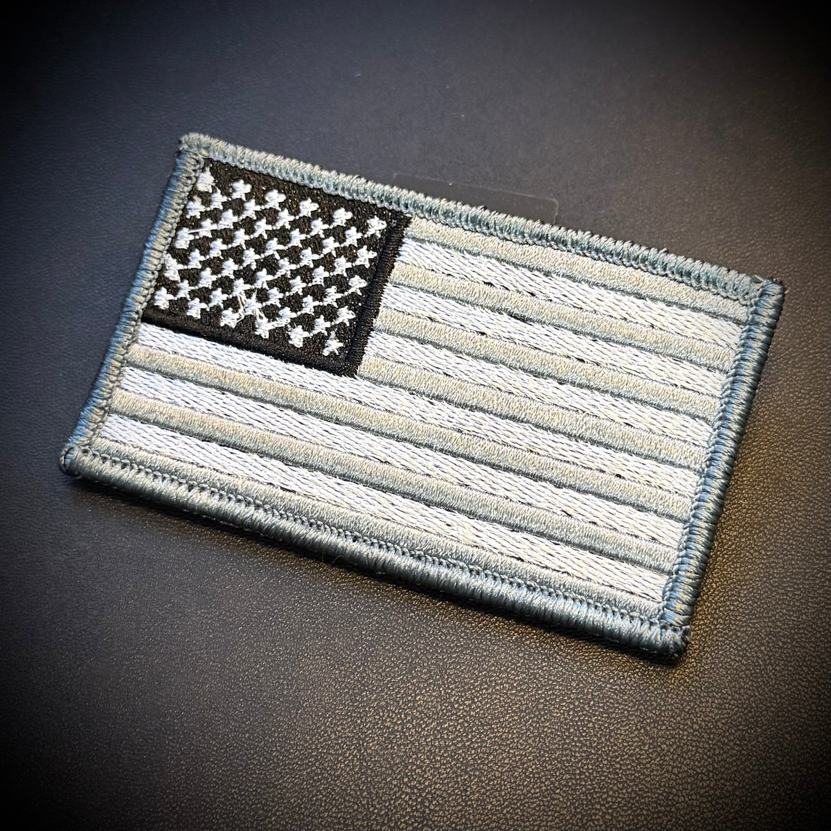 An embroidered tactical American flag patch in a monochrome black, white, and gray color scheme, displayed on a dark textured leather surface.