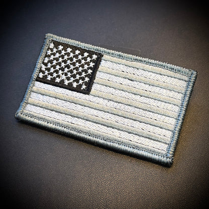 An embroidered tactical American flag patch in a monochrome black, white, and gray color scheme, displayed on a dark textured leather surface.