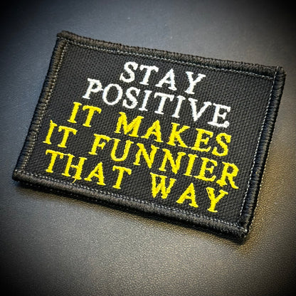 A rectangular black embroidered patch with the phrase 'STAY POSITIVE IT MAKES IT FUNNIER THAT WAY' in white and yellow lettering, shown on a dark textured background.