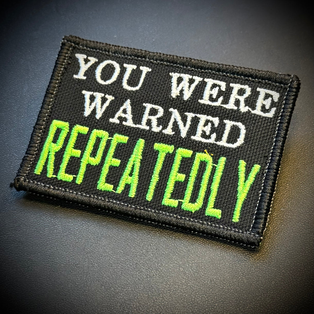 A rectangular embroidered patch with a black background and stitched text reading 'YOU WERE WARNED REPEATEDLY' in white and bright neon green, shown on a dark gray surface.