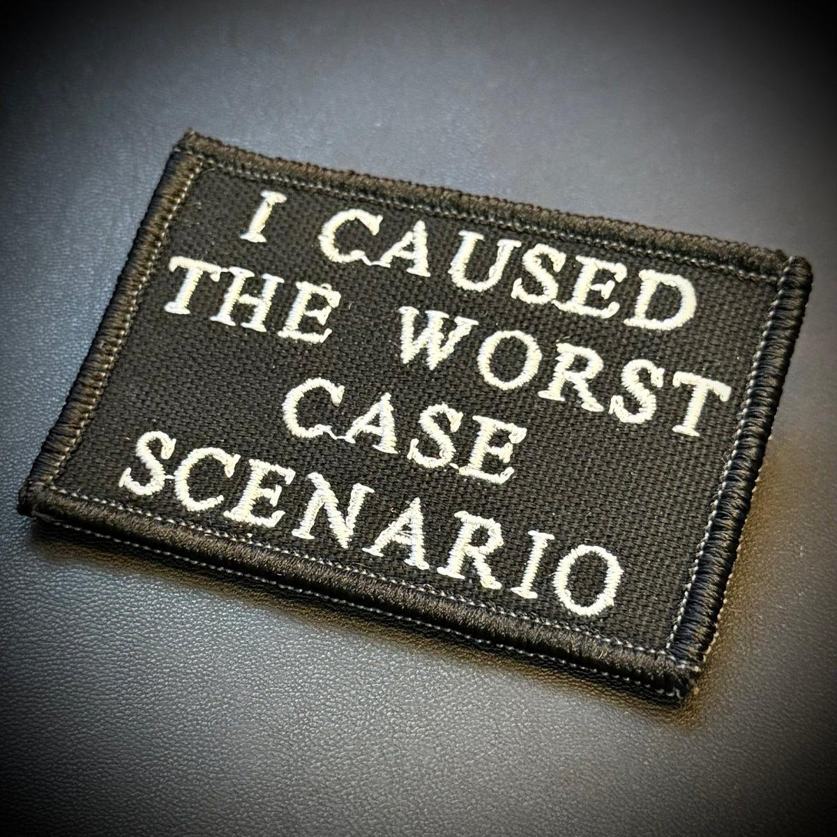 A rectangular embroidered patch with the phrase 'I CAUSED THE WORST CASE SCENARIO' in white lettering on a black background, photographed on a dark textured surface.