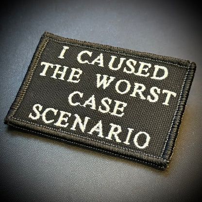 A rectangular embroidered patch with the phrase 'I CAUSED THE WORST CASE SCENARIO' in white lettering on a black background, photographed on a dark textured surface.
