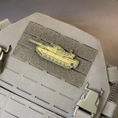 M1 Abrams Main Battle Tank laser-engraved synthetic morale patch: a gold-colored tank-shaped enamel pin attached to a gray hook-and-loop patch on a gray tactical backpack or gear panel.