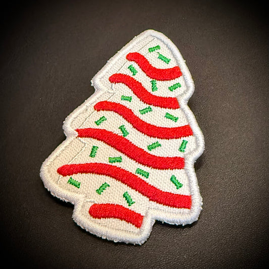 A Christmas tree-shaped embroidered patch with a white background, red wavy stripes, and small green accents, shown on a dark textured surface.