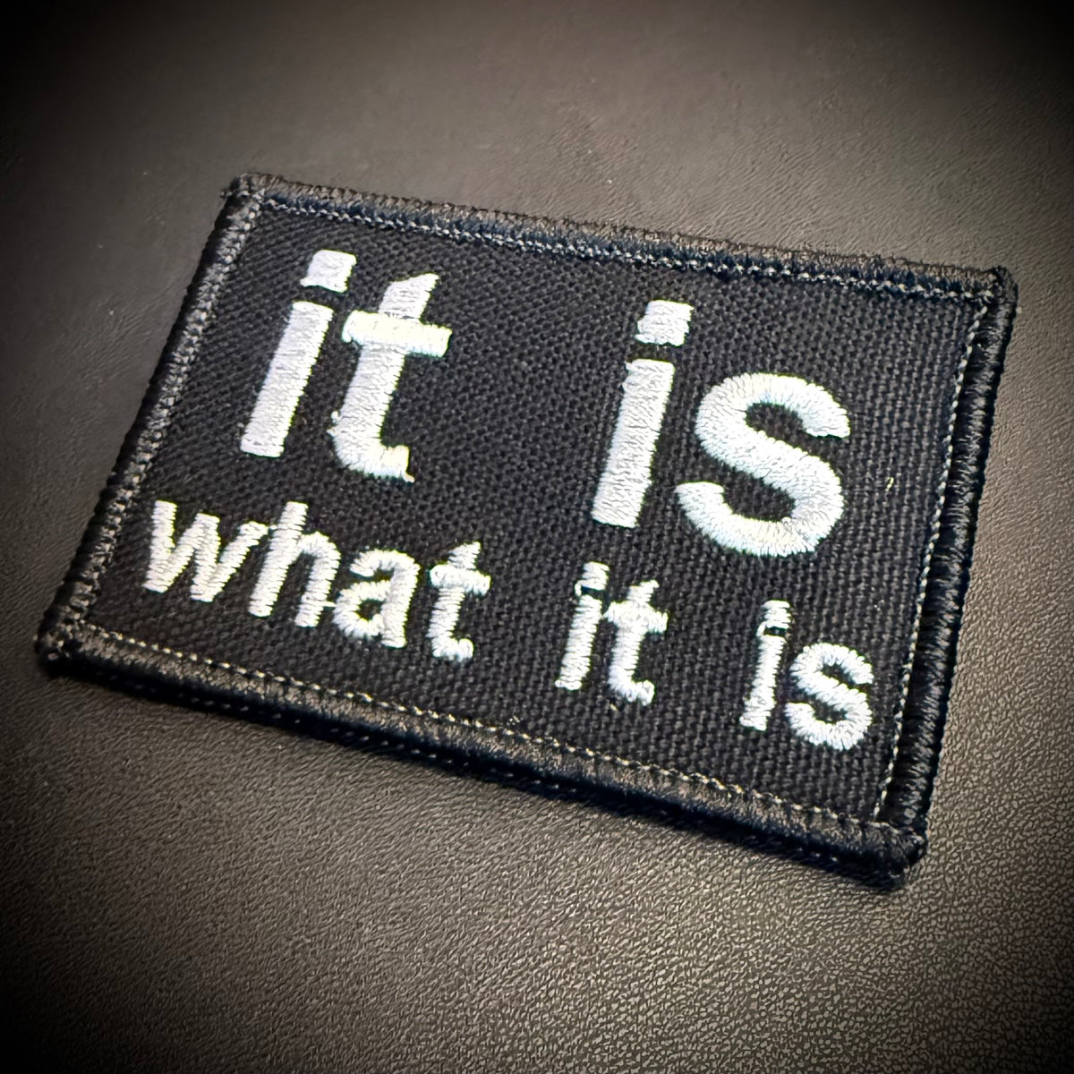 A rectangular black embroidered patch with white text reading 'it is what it is' on a dark textured background.