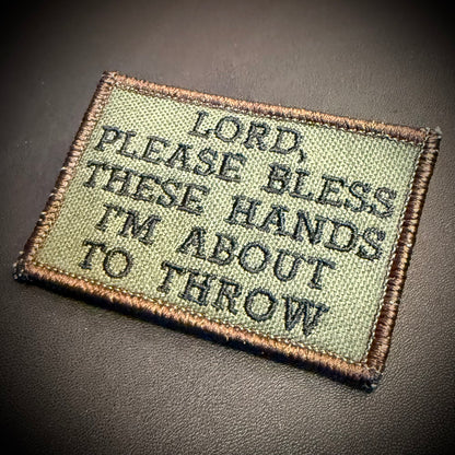 An embroidered rectangular patch with a tan border and olive green background, featuring the text 'LORD, PLEASE BLESS THESE HANDS I'M ABOUT TO THROW' in black lettering, shown on a dark textured surface.