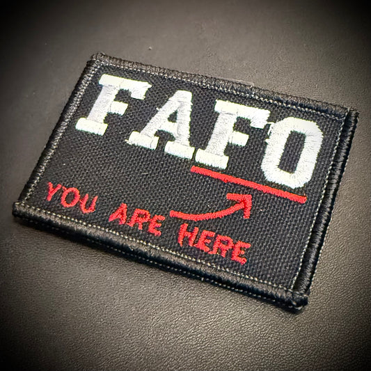 A rectangular embroidered patch with a black background, featuring the white distressed text 'FAFO' with a red underline and the red text 'YOU ARE HERE' with a red arrow, shown on a dark textured surface.