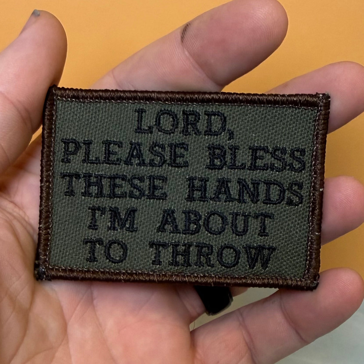 Lord, Bless These Hands I'm About To Throw embroidered morale patch: held in a hand, featuring the text 'LORD, PLEASE BLESS THESE HANDS I'M ABOUT TO THROW' in black on an olive drab background with a.
