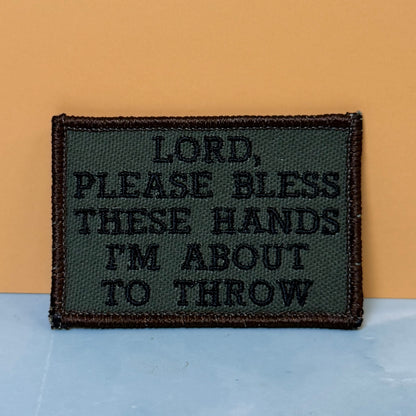 Lord, Bless These Hands I'm About To Throw embroidered morale patch: rectangular patch with the text 'LORD, PLEASE BLESS THESE HANDS I'M ABOUT TO THROW' in black thread on an olive green background, bordered in dark.