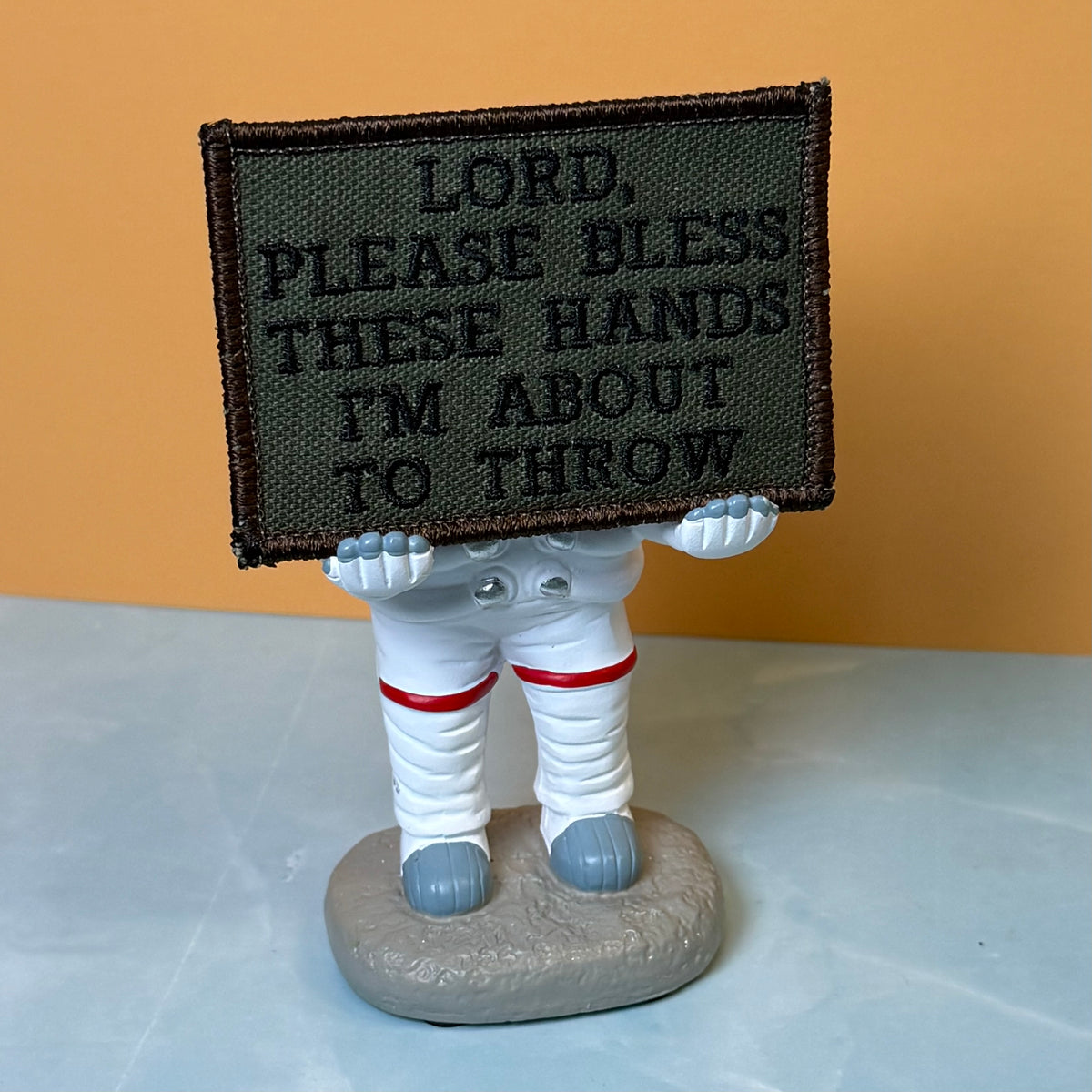 Lord, Bless These Hands I'm About To Throw embroidered morale patch: astronaut figurine holding a rectangular embroidered patch with the text 'LORD, PLEASE BLESS THESE HANDS I'M ABOUT TO THROW'. The astronaut is dressed.