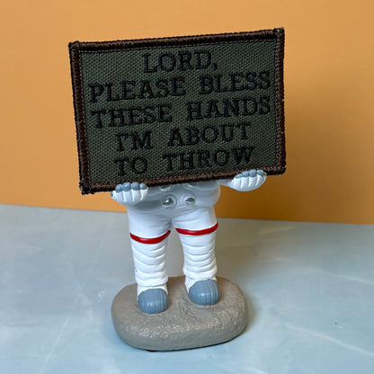 Lord, Bless These Hands I'm About To Throw embroidered morale patch: astronaut figurine holding a rectangular embroidered patch with the text 'LORD, PLEASE BLESS THESE HANDS I'M ABOUT TO THROW'. The astronaut is dressed.